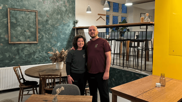 En kvinna med mörkt lockigt hår och en man i en vinröd t-shirt står tillsammans i en restaurang- eller caféinteriör. De står framför en mörkgrön, mönstrad fondvägg med en tom tavelram och ett runt träbord med stolar. Till höger bryter en starkt gul vägg a