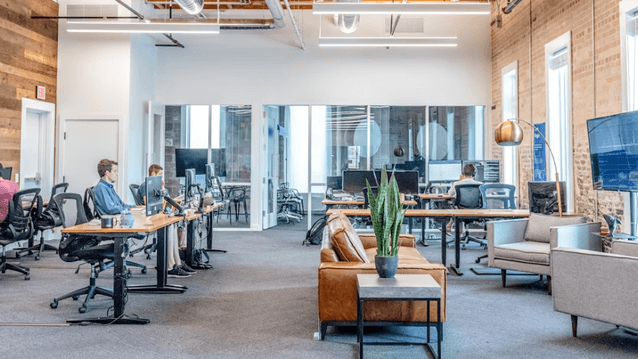 Ett ljust och öppet kontorslandskap i industristil med exponerade tegelväggar, trädetaljer och stora fönster. Personer arbetar vid långa skrivbord med datorskärmar, och i förgrunden syns en loungehörna med en brun skinnsoffa, fåtöljer och en stor grön väx