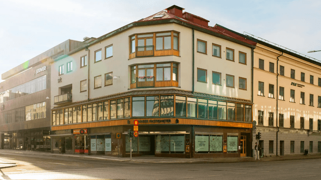 Hörnfastighet i tre våningar med ljus puts, trädetaljer och skyltfönster vid en korsning under en solig dag.