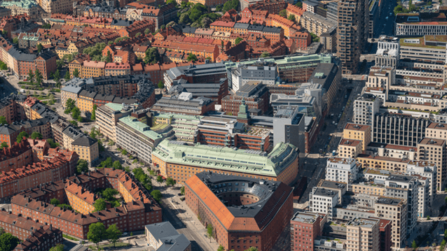 En flygbild över en tätbebyggd stadsmiljö med en unik triangelformad tegelbyggnad i centrum, omgiven av klassiska husrader och moderna höghus under en klarblå himmel.