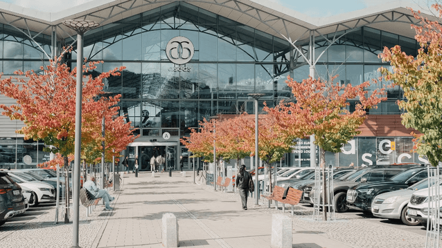 Entrén till köpcentret Asecs i Jönköping. Den stora glasfasaden speglar himlen och omges av parkerade bilar och en gångväg kantad av träd med färgstarka höstlöv. I förgrunden syns bänkar och människor som rör sig mot ingången.