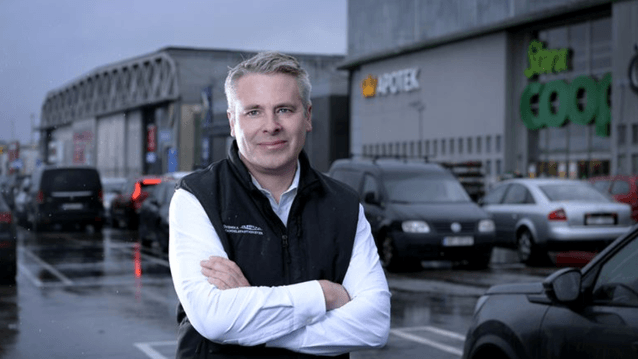 En man i vit skjorta och svart väst står med armarna i kors på en parkeringsplats framför en handelsplats. I den suddiga bakgrunden syns parkerade bilar och butikskedjor som "Kronans Apotek" och "Stora Coop". Det är en mulen dag och marken är blöt av regn