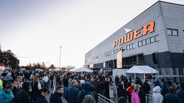 En stor folkmassa köar utanför en stor Power-elektronikbutik under dagsljus. Människor står tätt i fållor avgränsade med metallstängsel framför den grå butiksbyggnaden med den orangea Power-logotypen.