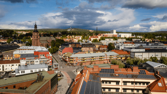 Flygfoto i fågelperspektiv över ett stadslandskap med kyrka, tegelbyggnader med solceller och skogsklädda höjder i bakgrunden under en dramatiskt molnig sky.