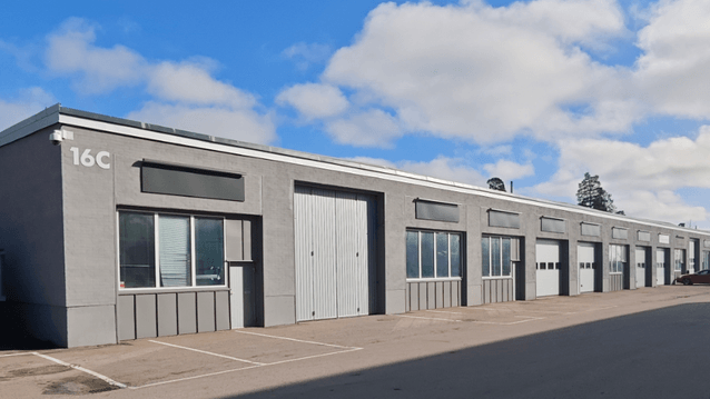 En lång, grå industrifastighet i en våning med flera sektioner under en blå himmel med lätta moln. Byggnaden har stora fönsterpartier, industriportar och en markerad husnummering "16C" på hörnet. Framför byggnaden finns en asfalterad yta med parkeringsmar
