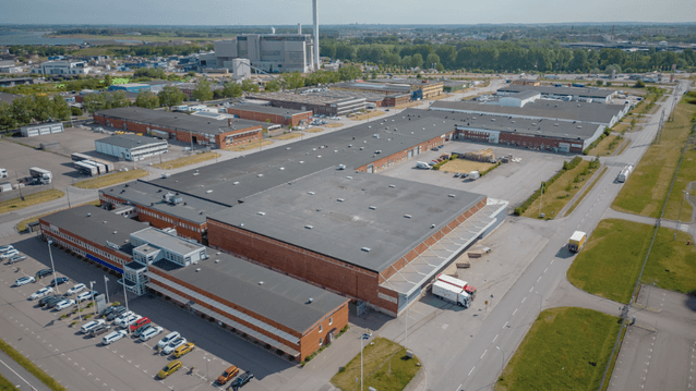 Flygbild över ett stort industriområde med flera tegelbyggnader med platta, mörka tak. En stor lastnings- och lossningsyta syns, och lastbilar står på en gata till höger. I bakgrunden syns en stor industrianläggning med en hög skorsten.