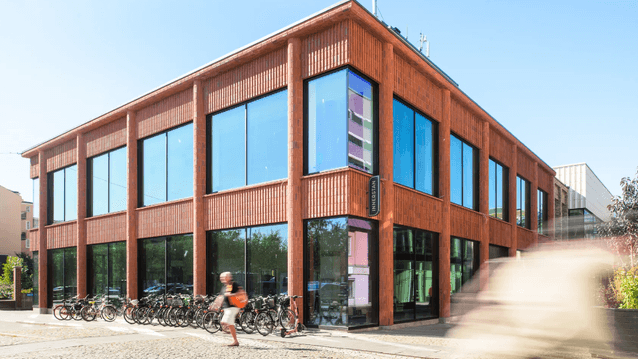 Hörnfasad på en tvåvåningsbyggnad i terracottafärg med stora glasskärmar, cykelparkering och en fotgängare i förgrunden.