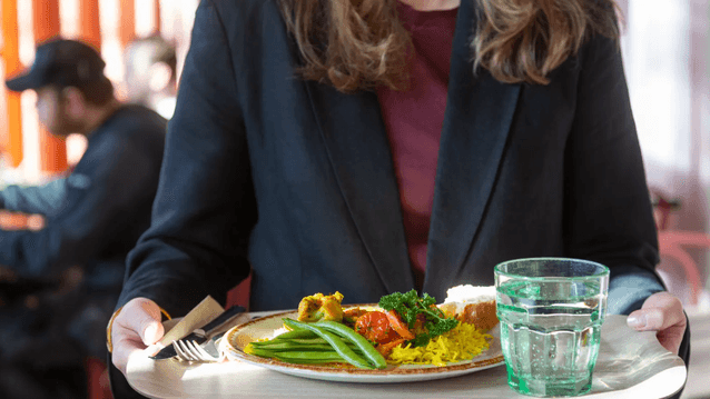 Närbild av en person som bär en bricka med en måltid bestående av ris, grönsaker samt ett glas vatten i en restaurangmiljö.