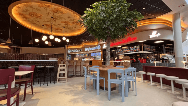Interiör från en modern food court eller restaurangtorg med sittplatser, ett runt bord byggt kring ett dekorativt träd, samt olika matställen i bakgrunden under stämningsfull belysning.