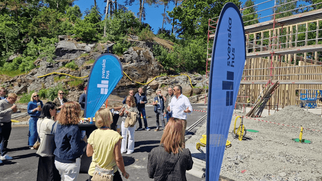 Byggstart och presentation vid en byggarbetsplats med företagsflaggor från Svenska Hus en solig dag.