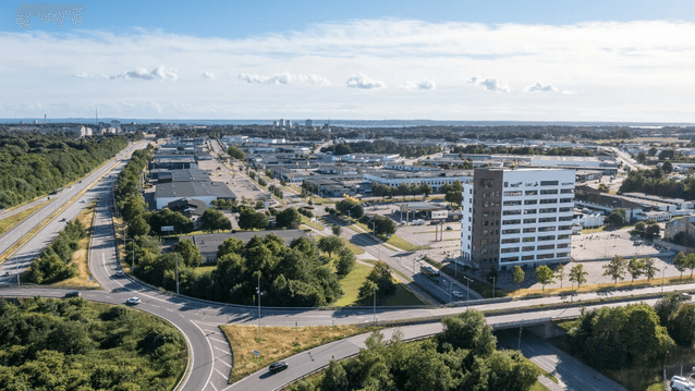 Högt flygfoto över ett industri- och handelsområde med en modern, ljus kontorsfastighet, trafikerade vägar och grönområden under en klarblå sky