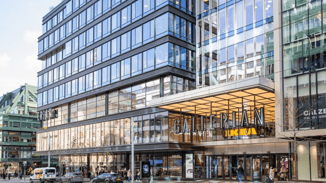 Exteriör av Gallerian i Stockholm med dess stora glasentré, där även skyltar för Ikea syns under det upplysta skärmtaket, och bilar som passerar på gatan utanför.
