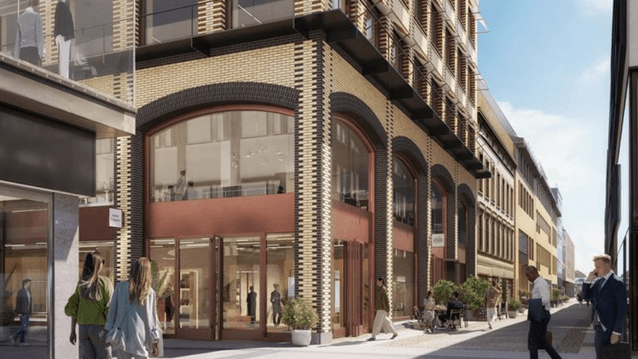 En digital visualisering av en modern stadsgata med en stor tegelbyggnad i fokus. Byggnaden har karaktäristiska rundade valv med stora glaspartier och butiker i gatuplan. Människor promenerar längs den ljusa gågatan och sitter på uteserveringar i solljuse