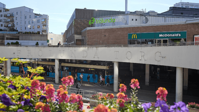 Kollektivtrafikknutpunkt vid Frölunda Torg köpcentrum i Göteborg, med en spårvagn vid hållplatsen och färgglada blommor i förgrunden.