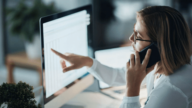 En kvinna med glasögon sitter vid ett skrivbord, talar i en smartphone och pekar på en datorskärm som visar ett kalkylark eller tabell.