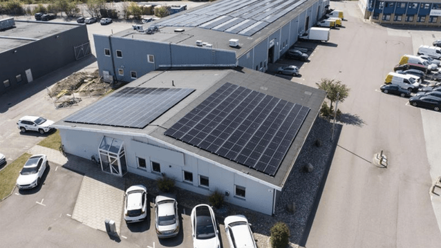 Flygfoto över en industrifastighet med stora solcellspaneler installerade på taken. Byggnaden omges av en parkeringsplats med bilar och ligger i anslutning till ett grönområde.