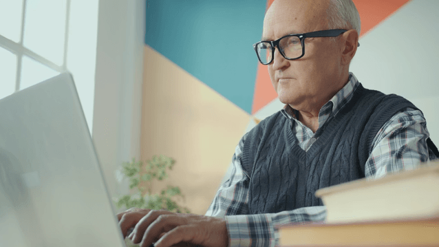 Närbild på en äldre man med glasögon och en stickad väst som sitter inomhus och fokuserat arbetar vid en bärbar dator. I bakgrunden syns ett fönster och en vägg med geometriska fält i turkos, rött och vitt. Till höger ligger en stapel böcker.