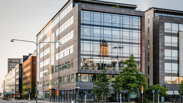 Exteriör av en modern kontorsbyggnad i glas och mörk sten vid Hjälmarekajen. I den stora glasfasaden reflekteras ett äldre torn, och framför byggnaden syns gröna träd, gatubelysning och en tom väg i mjukt dagsljus.