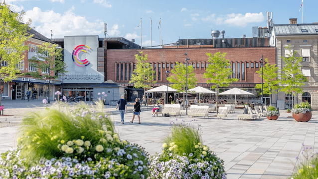Ett öppet stadstorg med ljus stenläggning framför köpcentret "Commerce". Byggnaden har en tegelfasad och en stor färgglad logotyp. På torget finns moderna sittmöbler, stora blomsterarrangemang i lila och gult, samt träd. Människor promenerar och sitter vi