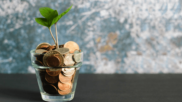 Ett litet grönt skott växer upp ur ett glas fyllt med olika mynt. Bilden symboliserar ekonomisk tillväxt eller sparande och har en suddig, melerad bakgrund i blåtoner.