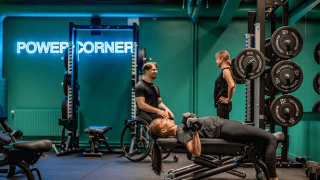 En kvinna gör hantelpress på en träningsbänk i ett gym med turkosa väggar och neonskylten "POWER CORNER".