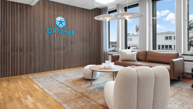 Interiör av ett modernt kontor med en loungehörna. En ribbad trävägg pryds av logotypen för "Optimal Advice". I rummet finns en brun lädersoffa, vita puffiga fåtöljer, ett runt soffbord på en stor mönstrad matta och stora fönster som släpper in dagsljus.