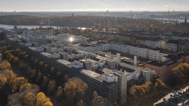 Flygfoto över ett stort sjukhus- eller universitetskomplex i en stad under hösten. Komplexet består av flera mörka, långa byggnader med platta tak och tekniska installationer, omgivet av träd med gulbruna löv. I bakgrunden syns Stockholms stadssiluett med