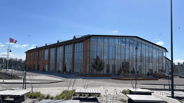 En stor industribyggnad i tegel med en modern glasfasad kallad Martinverket, omgiven av öppna ytor och bänkar under en klarblå himmel.