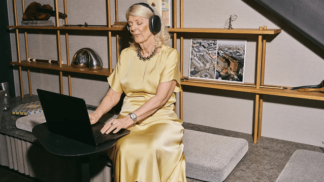 En kvinna i guldsidenklänning och stora hörlurar arbetar vid en bärbar dator. Hon sitter i en modern miljö med en dekorativ hylla i bakgrunden.