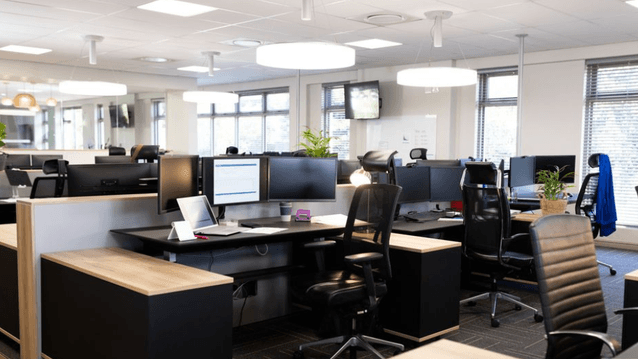 Ett ljust och modernt kontorslandskap med tomma arbetsplatser. Skrivborden är utrustade med dubbla bildskärmar, bärbara datorer och ergonomiska kontorsstolar. Stora runda taklampor ger ett mjukt ljus över lokalen.