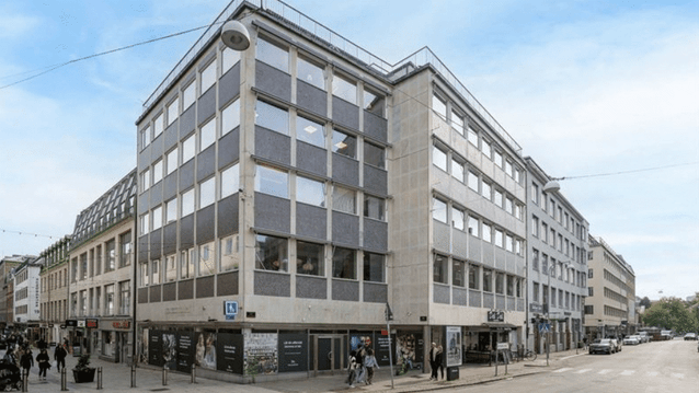 En hörnfastighet i flera våningar med grå och beige fasad i en stadsmiljö. Byggnaden ligger i korsningen mellan en gågata med flanörer och en bilväg, med butiksfönster på bottenvåningen.