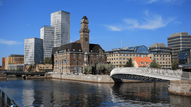 Utsikt över centrala Malmö med vattnet i förgrunden. Till vänster syns det moderna höghuset Malmö Live och andra kontorsbyggnader, medan den gamla tegelbyggnaden Malmö Börshus står i centrum med sitt torn. En vit gångbro sträcker sig över kanalen, och him