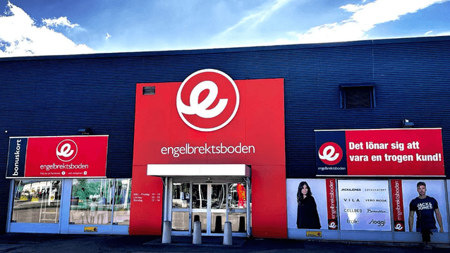 Exteriörbild i ögonhöjd över entrén till klädbutiken Engelbrektsboden med röda fasadskyltar och varumärkesreklam.