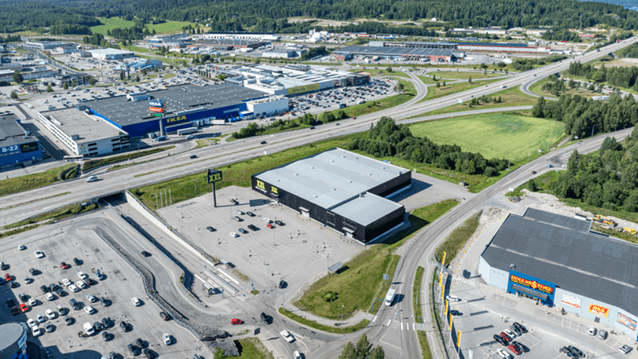 En flygbild över ett handelsområde med stora butikskedjor som IKEA, XXL och Dollarstore, omgivet av motorvägar, parkeringsplatser och skogsklädda kullar.