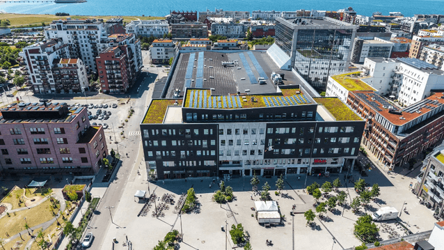 Flygfoto över ett torg och stadsområde med en stor mörk fastighet utrustad med solpaneler och växttak vid havet.