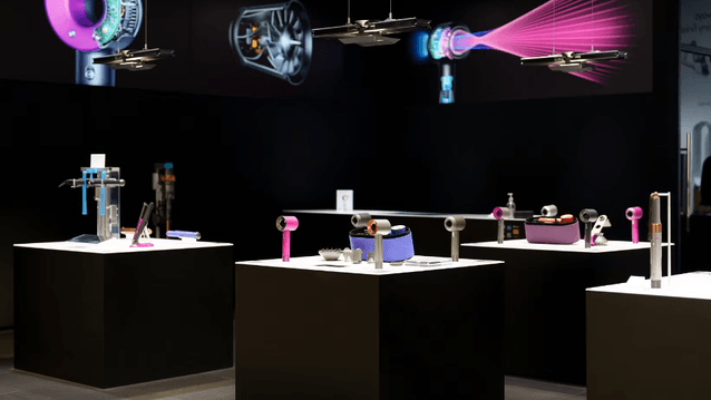En stilren produktutställning i en mörk butikslokal där olika Dyson-hårfönar och stylingverktyg presenteras på svarta podier. I bakgrunden syns stora belysta skärmar som visar tekniska sprängskisser och illustrationer av luftflöden i lila och blått. Produ