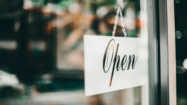 En närbild av en vit skylt med texten "Open" i kursiv stil, som hänger i ett fönster eller en glasdörr. Skylten hänger i en metallkedja som fästs med en sugpropp. Bakgrunden är suddig och visar gröna och bruna nyanser,
