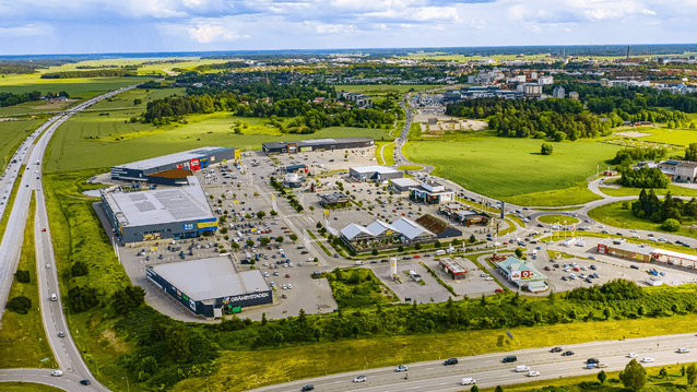 Flygfoto över handelsområdet Gränbystaden med stora butiksfastigheter, parkeringsplatser, vägar och åkerlandskap i bakgrunden.