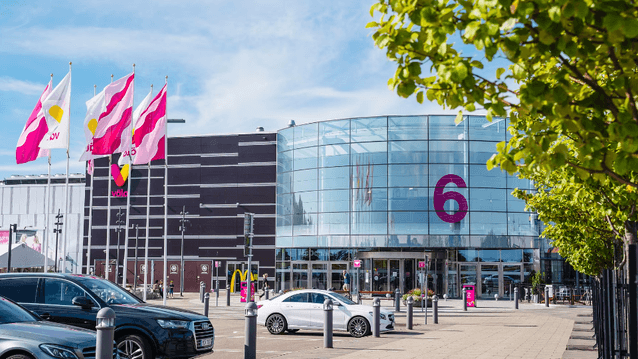 Väla köpcentrums entré nummer 6 med en stor rundad glasfasad, flaggor som vajar i vinden och parkerade bilar i förgrunden.