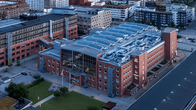 Den stora tegelbyggnaden har ett modernt glaskonstruerat mittparti och taket är täckt med solcellspaneler. Byggnaden ligger i ett urbant område nära hamnen med flertalet bostadshus i bakgrunden.