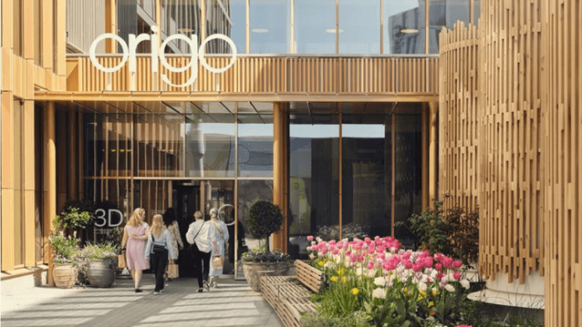 En modern byggnad med namnet "Origo" ovanför entrén. Fasaden är klädd i ljusa träribbor och stora glaspartier. I förgrunden syns en stenlagd gångväg kantad av blommande rosa och vita tulpaner, och en grupp människor är på väg in i byggnaden.