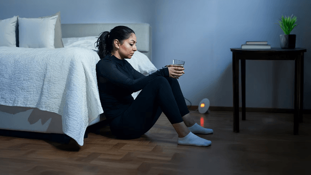 En mörk inomhusbild visar en kvinna i svarta, åtsittande kläder sittande på ett trägolv med korslagda ben, lutad mot en säng med vitt täcke. Hon håller ett glas vatten och ser tankfull ut. I det svagt upplysta rummet syns ett nattduksbord med böcker och e