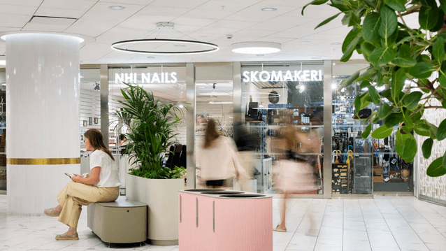 Ljus galleriagång med butikerna NHI Nails och Skomakeri, en sittande kvinna samt en rosa källsorteringsmöbel.
