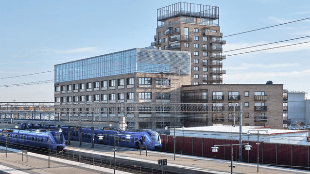 Blå pendeltåg vid en tågstation framför en modern tegelbyggnad med stora glaspartier och en hög huskropp.