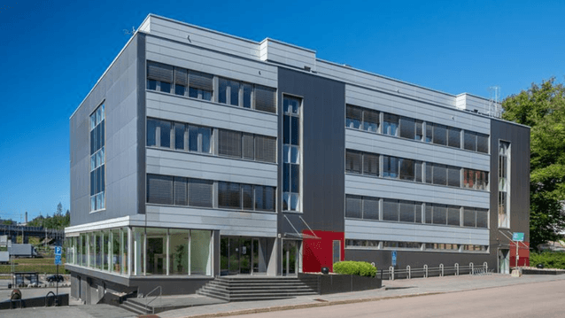Modern kontorsbyggnad i fyra våningar med grå fasadpaneler, röda accentdetaljer och entrétrappa mot gatan under en klarblå sky.