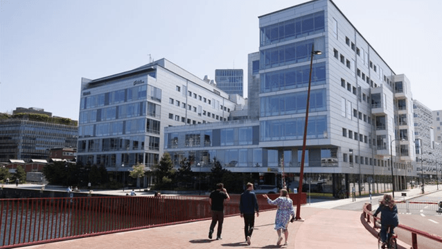 En röd gång- och cykelbro där människor passerar framför ett stort, modernt kontorskomplex med glasfasad.