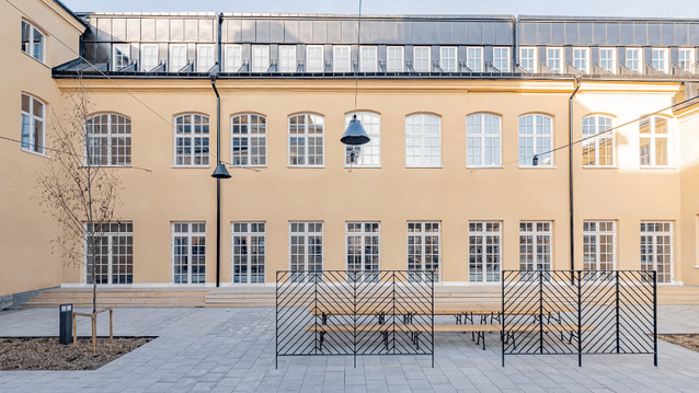 En stenlagd innergård framför en klassisk, ljusgul byggnad med regelbundna fönsterrader, bänkar och svarta metallskärmar.