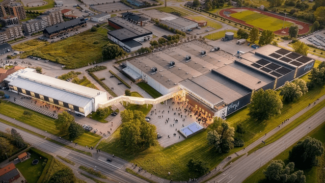 En flygbild över Plejds huvudkontor och produktionsanläggning i Mölndal. Den moderna industrifastigheten har stora lagerhallar och en vit kontorsbyggnad som är sammanlänkad via en inglasad gångbro. Framför byggnaden syns en öppen samlingsplats med människ