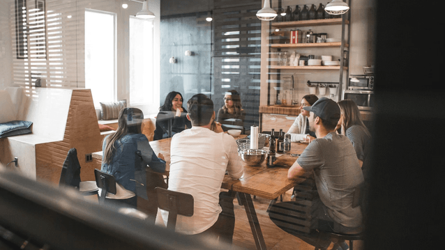 En grupp människor sitter samlade runt ett långt träbord i en avslappnad kontorsmiljö eller fikarum. De samtalar i ett ljust rum med industriell känsla, där en öppen hylla med porslin och stora fönster syns i bakgrunden.