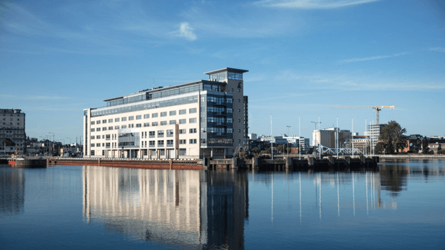 Ett modernt, ljusfärgat kontorshus vid vattnet reflekteras tydligt i den stilla vattenytan. Huset har flera våningar och glaspartier, och bakom byggnaden syns en stadsmiljö med fler hus, en byggkran och träd. Himlen är klarblå med några tunna moln.